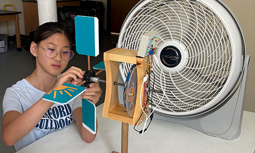 A boy and a girl work on a turbine in front of a fan.