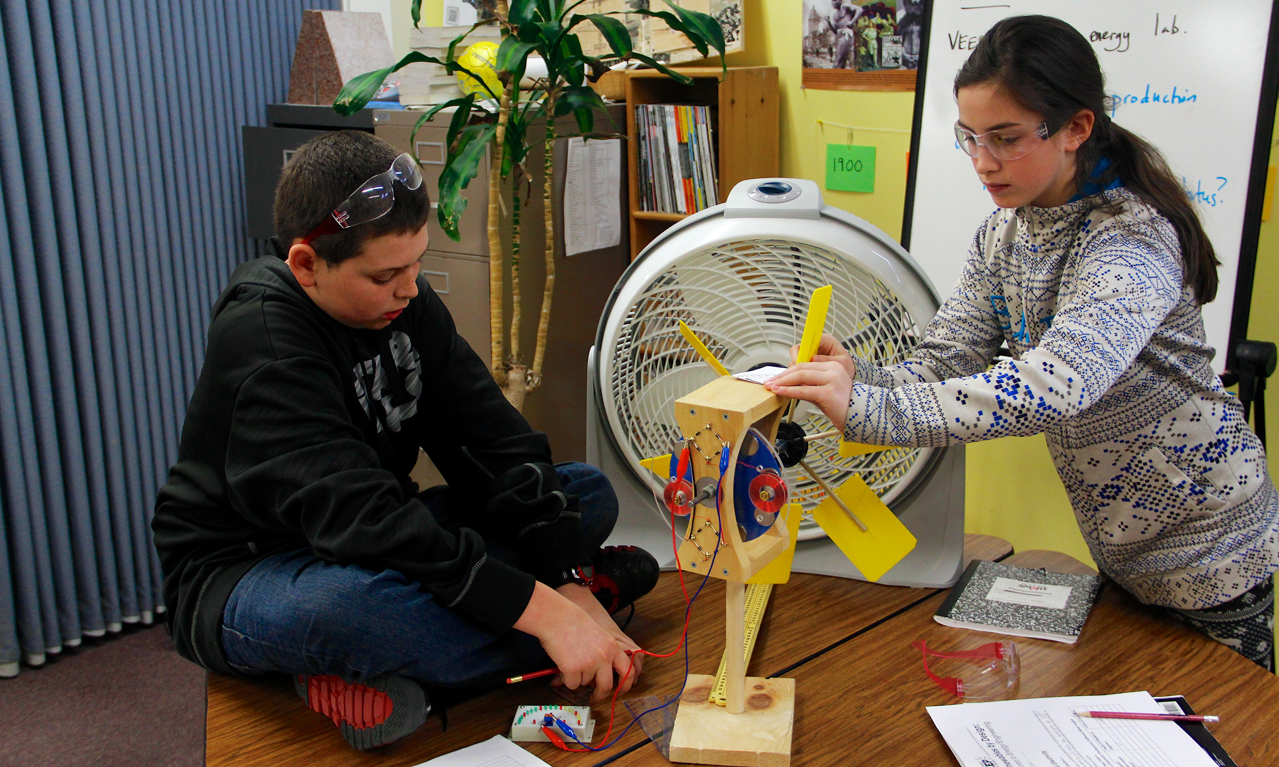 A boy and a girl work on a turbine in front of a fan.